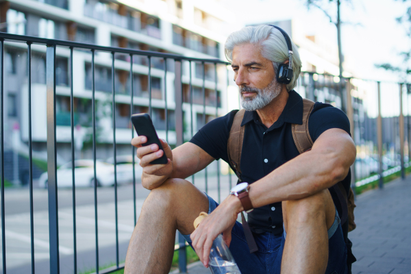 Portrait of handsome mature man with headphones sitting outdoors in city, using smartphone.