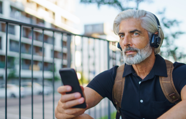 Portrait of handsome mature man with headphones sitting outdoors in city, using smartphone.