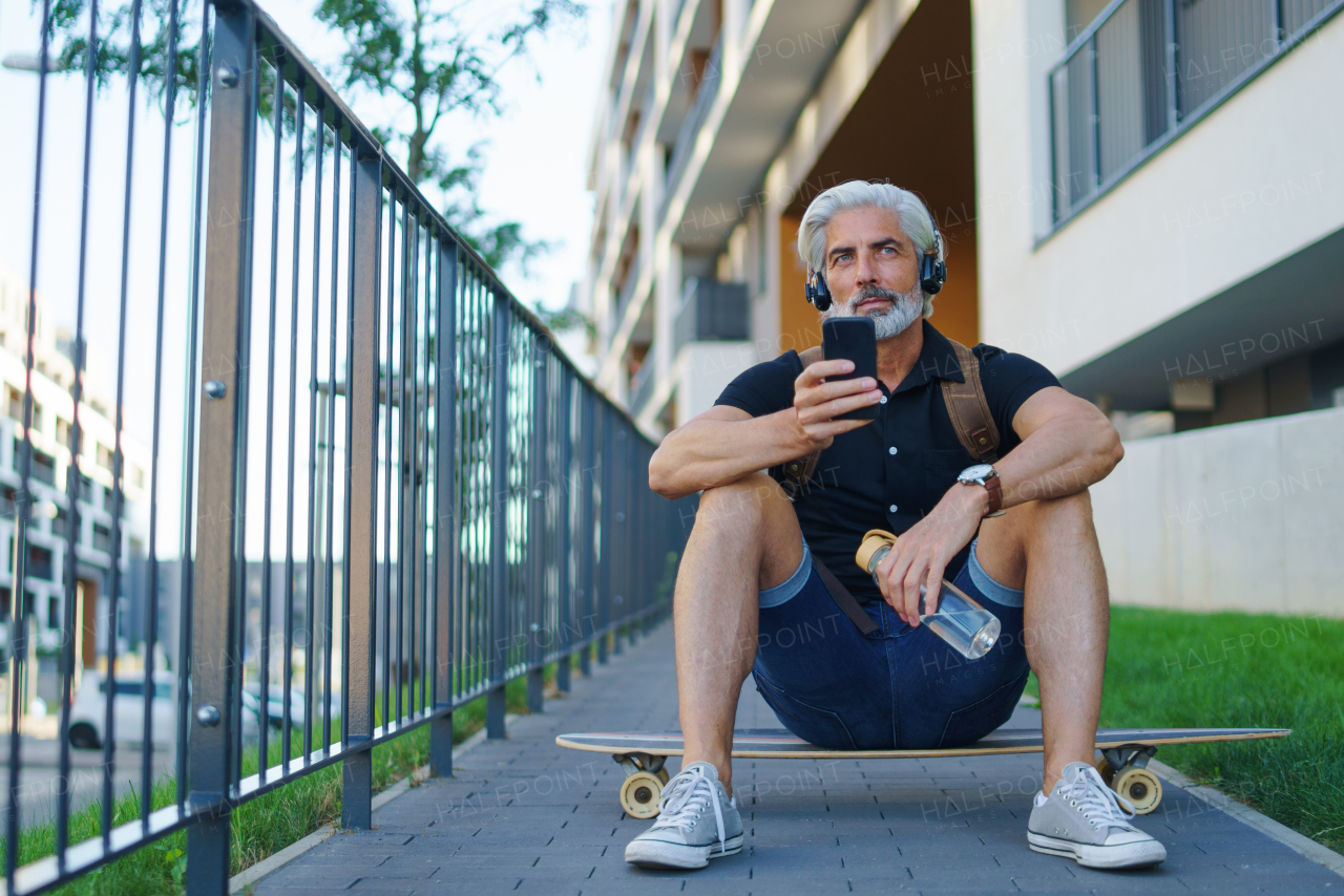 Portrait of handsome mature man with headphones sitting outdoors in city, using smartphone.