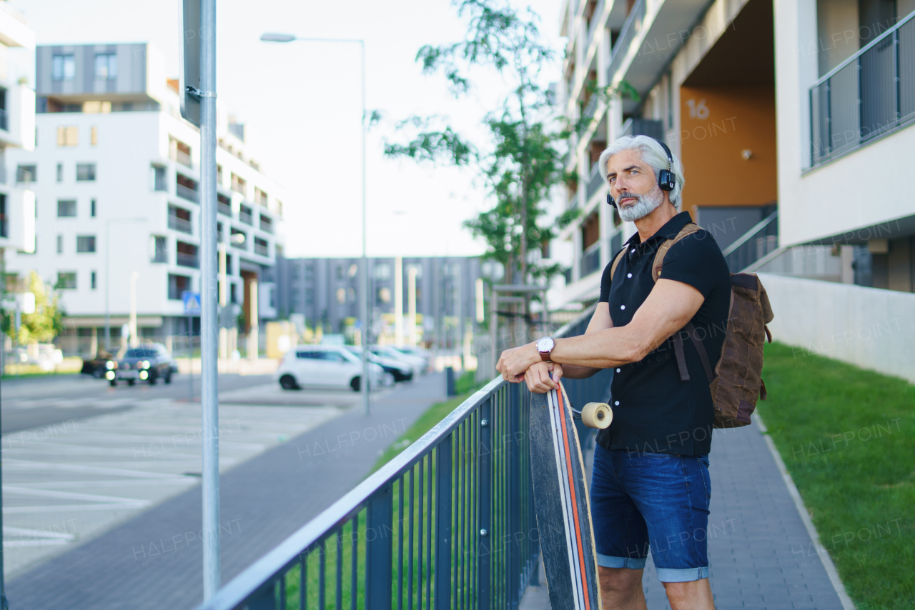 Side view portrait of mature man with skateboard and headphones outdoors in city, going back to work.