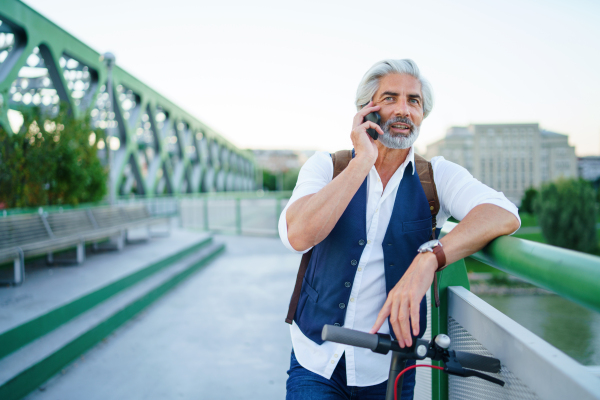 Portrait of mature man with electric scooter standing outdoors in city, using smartphone.