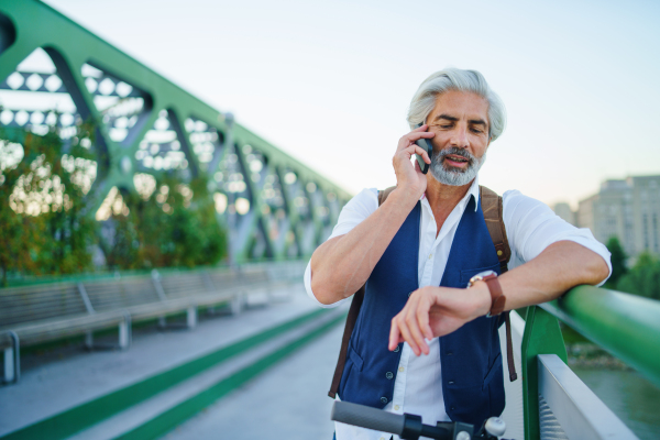 Portrait of mature man with electric scooter standing outdoors in city, using smartphone.