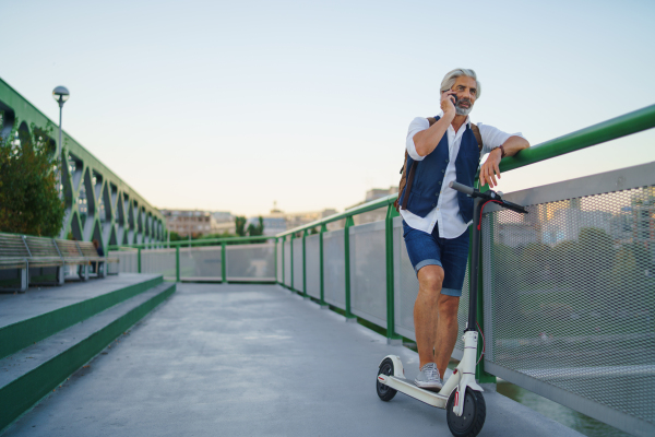 Portrait of mature man with electric scooter standing outdoors in city, using smartphone.