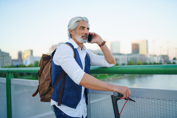 Portrait of mature man with electric scooter standing outdoors in city, using smartphone.