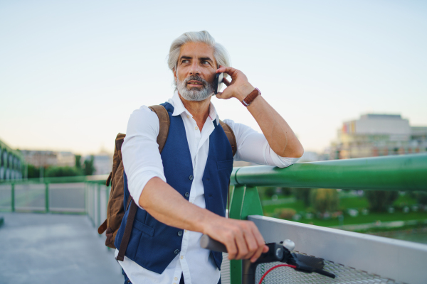 Portrait of mature man with electric scooter standing outdoors in city, using smartphone.