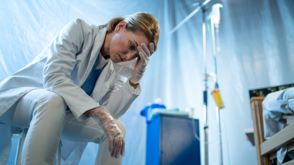 Tired and frustrated doctor sitting by patient in bed in hospital, coronavirus concept.