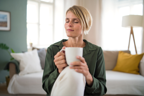 Front view portrait of woman meditating indoors in office, holding cup of tea.