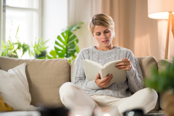 Front view portrait of woman relaxing and reading book indoors at home, mental health care concept.