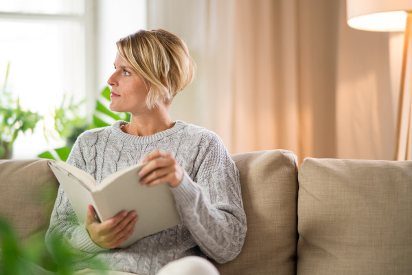 Front view portrait of woman relaxing and reading book indoors at home, mental health care concept.