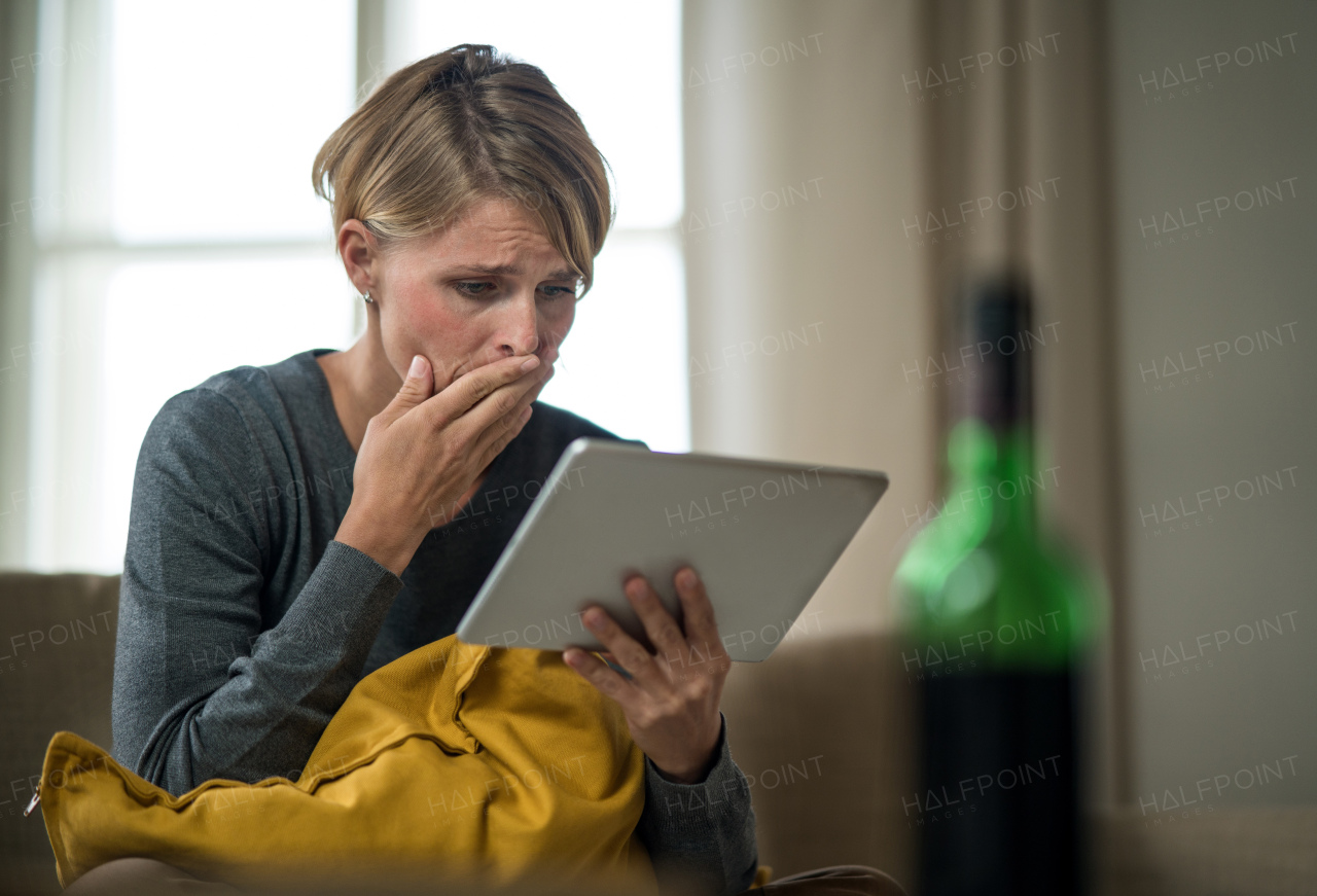 Worried woman with tablet indoors on sofa at home feeling stressed, mental health concept.