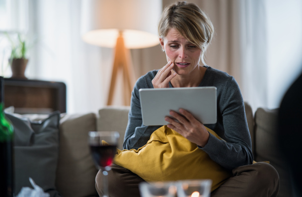 Worried woman with tablet indoors on sofa at home feeling stressed, mental health concept.