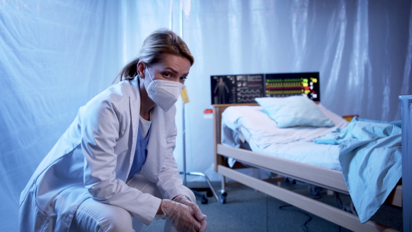 Tired and frustrated doctor sitting by patient in bed in hospital, coronavirus concept.