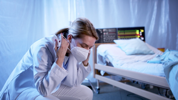 Tired and frustrated doctor sitting by patient in bed in hospital, looking at camera. Coronavirus concept.