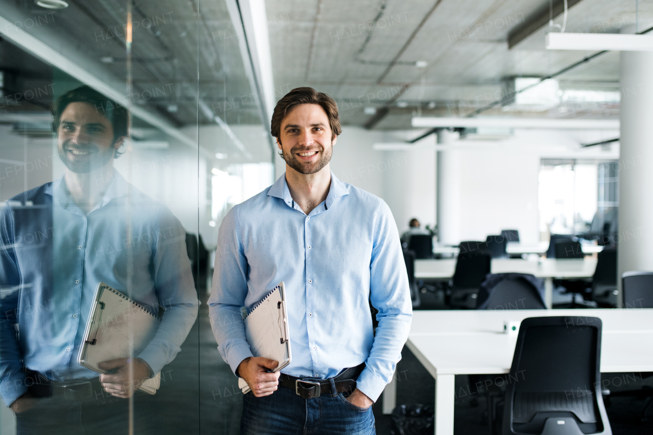 A portrait of young businessman standing in an office, looking at camera.