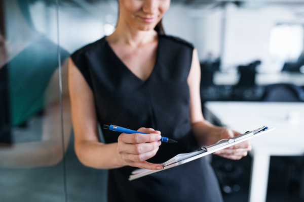 A midsection of young businesswoman with clipboard in an office, standing.