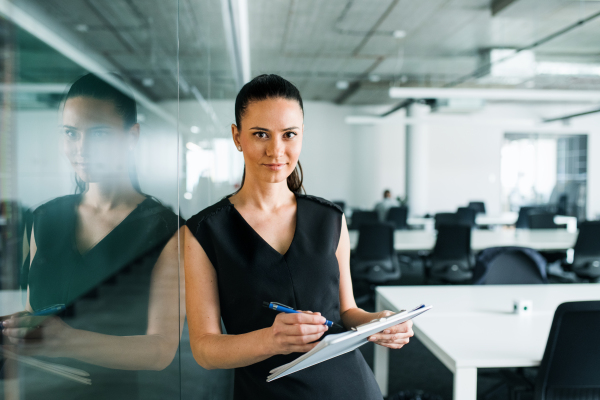 A front view of young businesswoman with clipboard in an office, standing.