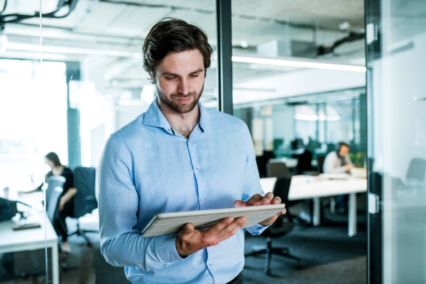 A portrait of young businessman with tablet standing in an office, working.