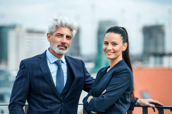 A mature businessman and young businesswoman standing on a terrace, looking at camera.