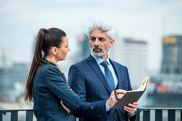 A mature businessman and young businesswoman with diary standing outdoors on a terrace, working.