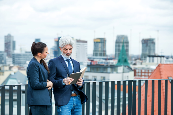 A mature businessman and young businesswoman with diary standing outdoors on a terrace, working.