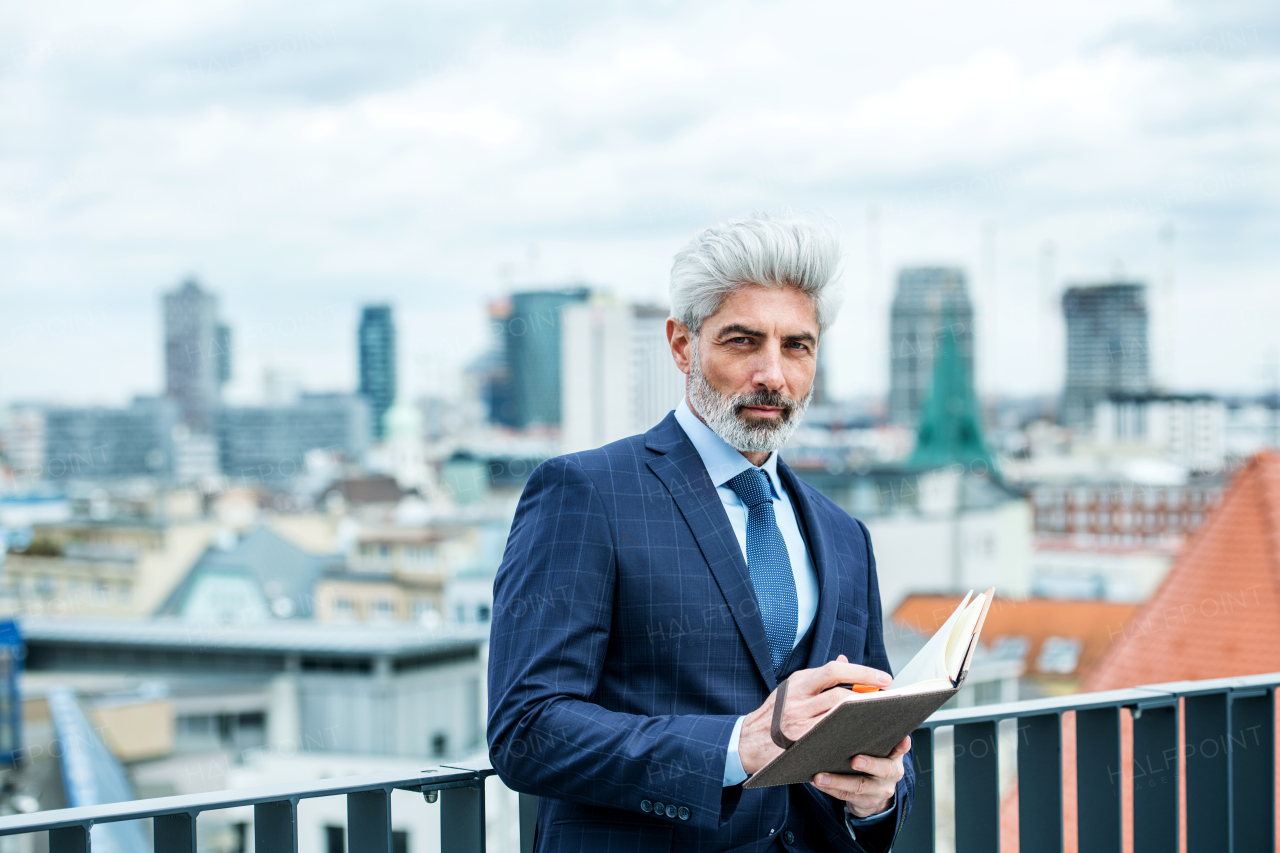 A mature businessman with diary standing on a terrace outside office, writing and planning.