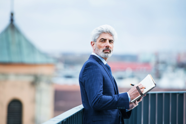 A mature businessman with diary standing on a terrace outside office, writing and planning.