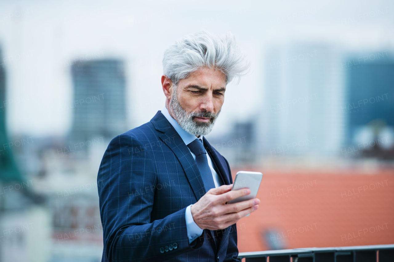 A content mature businessman standing on a terrace, using smartphone.