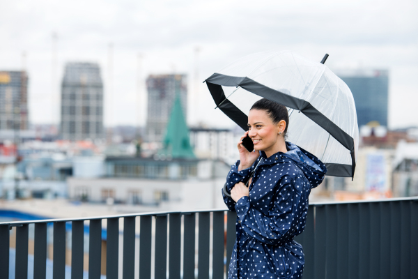 A young woman with smartphone and umbrella and anorak standing on a terrace, making a phone call.