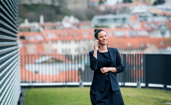 A front view of young businesswoman with smartphone standing on a terrace, making a phone call.