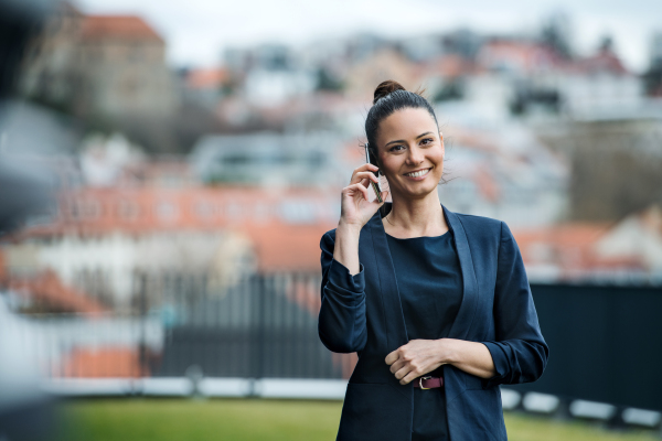 A front view of young businesswoman with smartphone standing on a terrace, making a phone call.