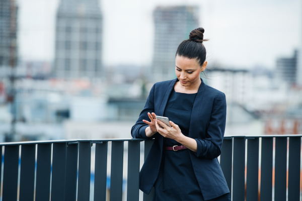 A young businesswoman standing outdoors on a terrace, using smartphone.