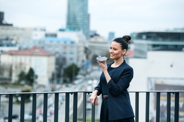 A front view of young businesswoman with smartphone standing on a terrace, making a phone call.