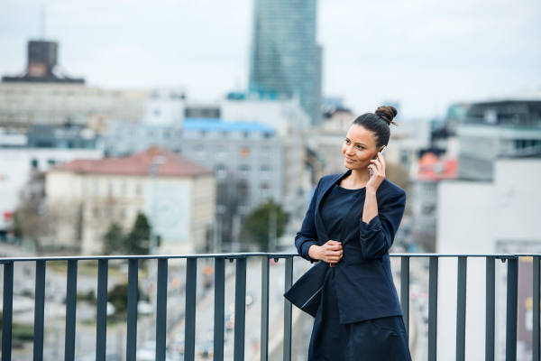 A front view of young businesswoman with smartphone standing on a terrace, making a phone call.