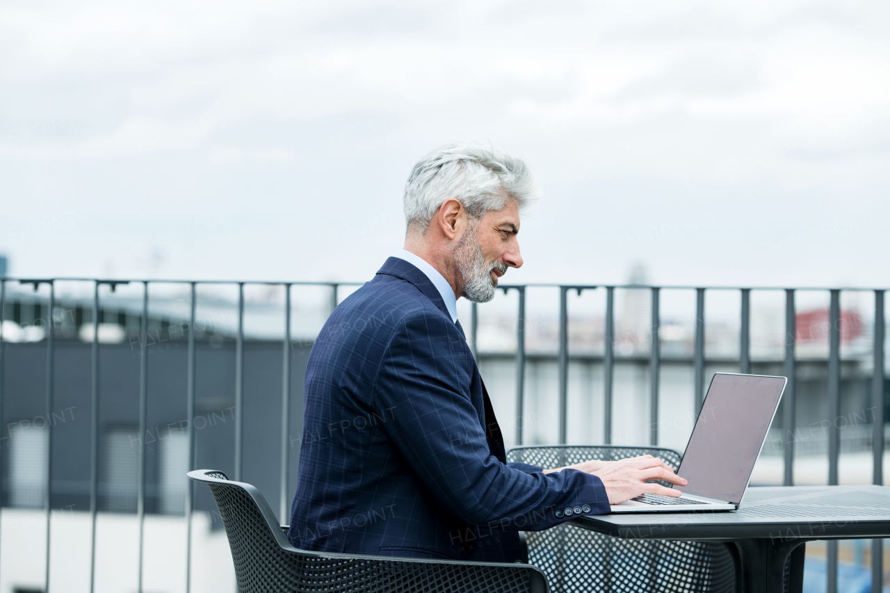 A mature businessman with laptop sitting at the table on a terrace, working.