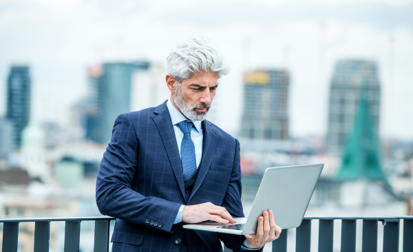 A mature businessman with laptop standing on a terrace outside office, working.