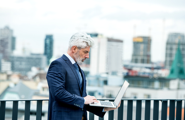 A mature businessman with laptop standing on a terrace outside office, working.