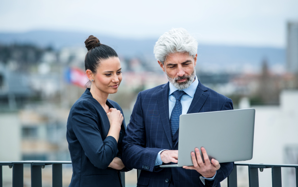 A mature businessman and young businesswoman with laptop standing outdoors on a terrace, working.