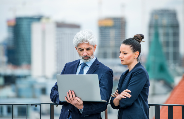 A mature businessman and young businesswoman with laptop standing outdoors on a terrace, working.