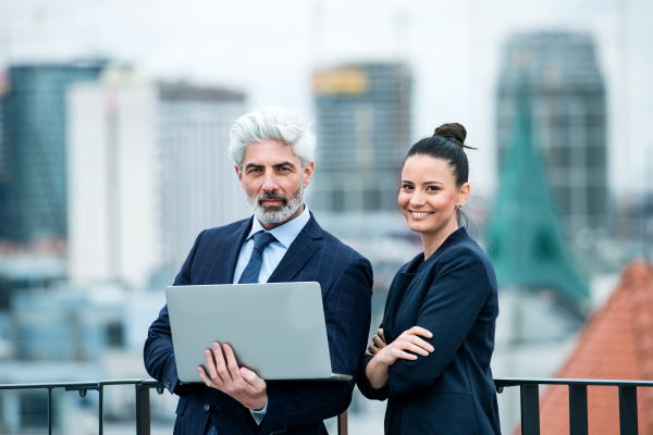 A mature businessman and young businesswoman with laptop standing outdoors on a terrace, working.