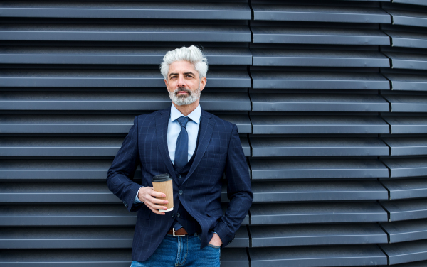 A mature businessman standing outdoors, holding paper cup with coffee. Copy space.