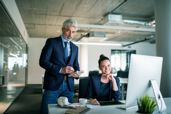 Two businesspeople with computer sitting in an office at desk, looking at camera.