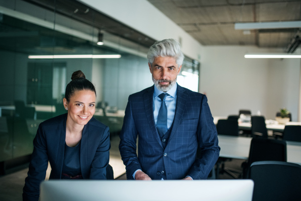 Two businesspeople with a computer standing in an office at desk, looking at camera.