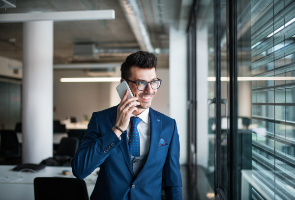 A portrait of young businessman with smartphone standing in an office, making phone call.