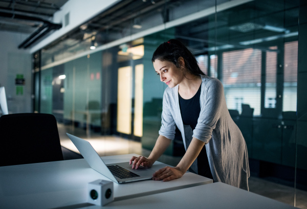 Side view of young businesswoman with computer standing at the desk in an office, working.