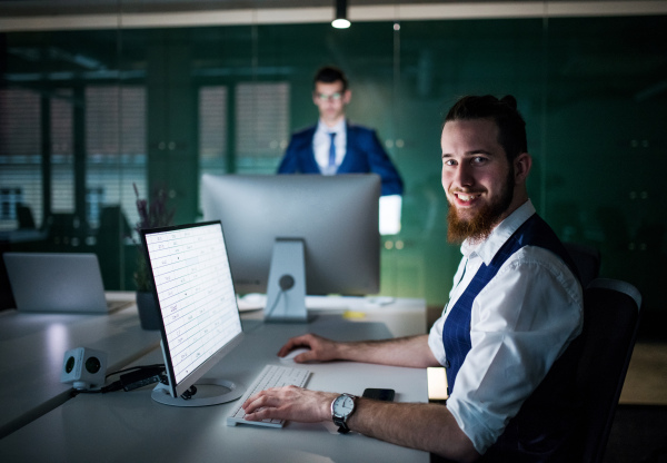 A portrait of young businessman with laptop sitting in an office, looking at camera.