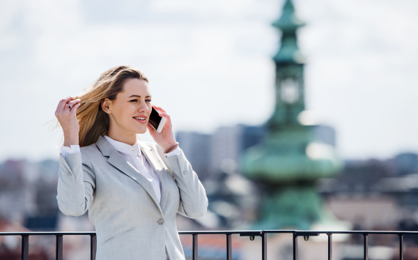 A young businesswoman with smartphone and laptop standing on a terrace, making a phone call when working.