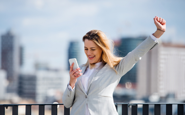 A young businesswoman with smartphone standing on a terrace outside office, expressing excitement.