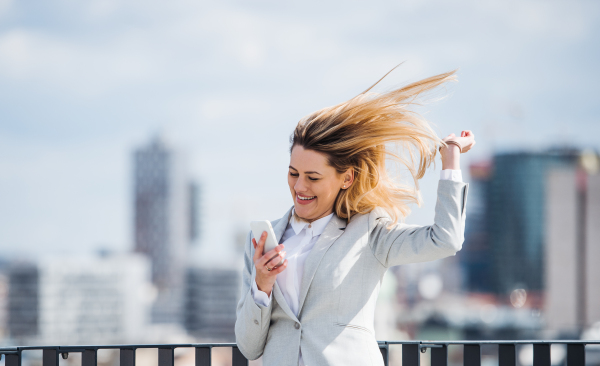 A young businesswoman with smartphone standing on a terrace outside office, expressing excitement.