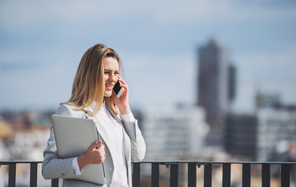 A young businesswoman with smartphone and laptop standing on a terrace, making a phone call when working.