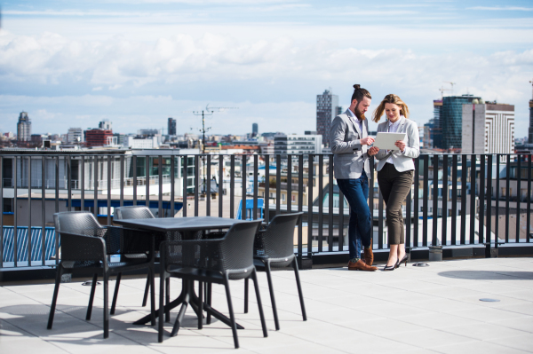 Two cheerful young business people with tablet standing on a terrace outside office, working.